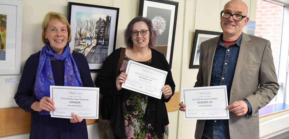 Mary Fawcett, Joanne Tudor and Fred Fieber holding certificates in front of the exhibition