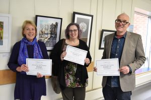 Mary Fawcett, Joanne Tudor and Fred Fieber holding certificates in front of the exhibition