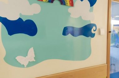 Butterfly rainbow print on wall with cut out shaoes of sky and clouds and a butterfly ready for messages from parents and staff