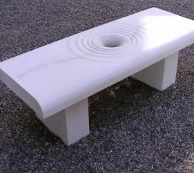 Carved stone bench with hole in centre and concentric circles like ripples, line of poetry