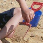 boy's hand holding red spade full of sand, blue bucket behind